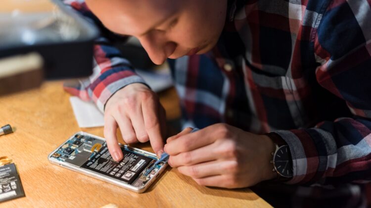 An individual is working on the inside of a phone, using a tool to manipulate electronic components. A wooden table is visible, with spare parts next to it.